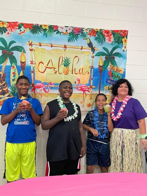 Students with Tricia with shaved ice drinks in front of a Hawaiian themed poster.