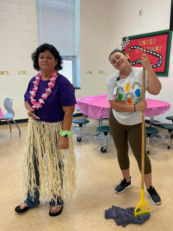 Tricia wearing a lei and grass skirt with Michaela holding a mop