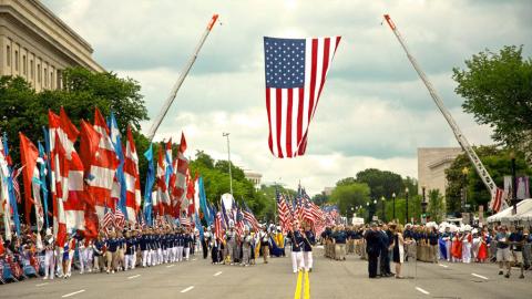 Memorial Day parade on a street with a giant U.S. flag hanging from cranes.
